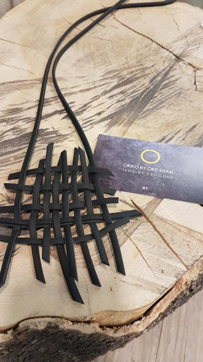 May include: Black leather necklace with a woven design. The necklace is on a wooden surface. The card reads "Oriko by Ort Efan Jewelry Designer".