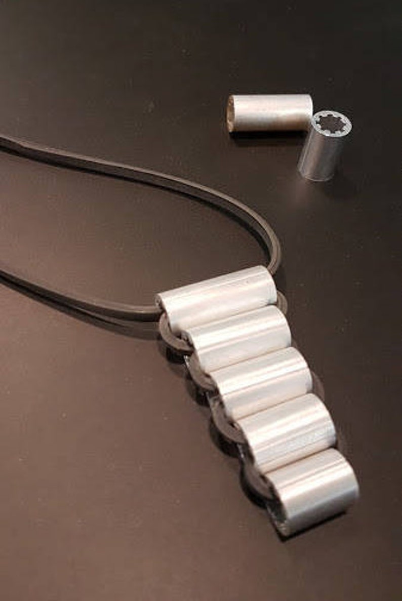May include: A silver necklace with a chain made of multiple connected cylinders. The necklace is on a black surface.