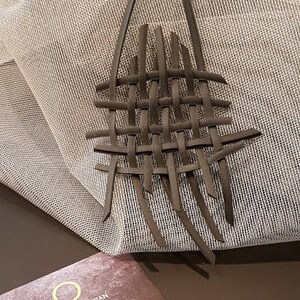 May include: A brown leather necklace with a woven design. The necklace is on a white fabric background. The text "ORIKO BY ORIT ETAN JEWELRY DESIGNER" is on a brown card.