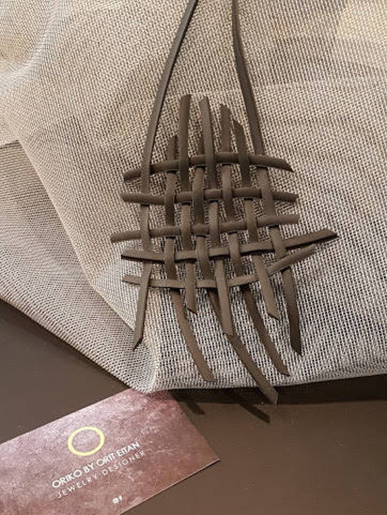 May include: A brown leather necklace with a woven design. The necklace is on a white fabric background. The text "ORIKO BY ORIT ETAN JEWELRY DESIGNER" is on a brown card.