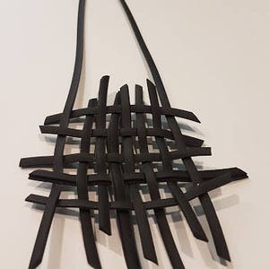 May include: A black woven necklace with a long, thin chain. The necklace is made of multiple strands of black material that are woven together to create a lattice pattern.