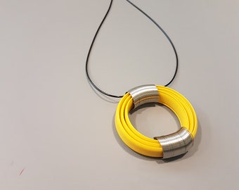 Puede incluir: Un collar amarillo y plateado con un colgante circular. El colgante está hecho de varias capas de material amarillo con una banda plateada envuelta alrededor del centro.