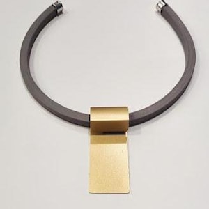Peut inclure: Un collier moderne avec un bandeau arrondi gris foncé et des extrémités en métal argenté. Un pendentif rectangulaire de couleur or est suspendu au centre. Le collier est présenté sur un fond gris clair.