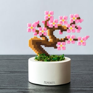 Op de afbeelding: Een miniatuur kersenbloesemboom gemaakt van roze en bruine perler kralen, die in een witte pot met groen mos staat. De pot heeft de tekst "PXLPRINCESS" op de bodem.