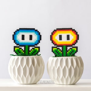 May include: Two pixel art flower pots, one blue and one orange, in white ceramic pots with a textured design. The flowers are made of perler beads and resemble the power-up flowers from the Super Mario Bros. video game.