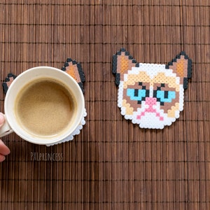 May include: A white ceramic coffee mug with brown coffee inside, sitting on a brown and white pixel art coaster of a grumpy cat with blue eyes. The coaster is on a brown bamboo mat.