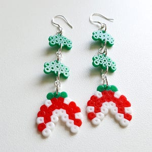 Puede incluir: Un par de pendientes colgantes con un dise&ntilde;o de cuentas verdes y rojas. Los pendientes presentan tres cuentas verdes en forma de tr&eacute;bol y una cuenta roja y blanca en forma de semic&iacute;rculo.
