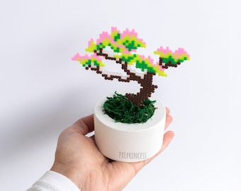 Może przedstawiać: Mała, biała ceramiczna doniczka z zielonym mchem i różowo-zielonym drzewem bonsai w pikselach. Doniczka ma napis "PXLPRINCESS" na spodzie.