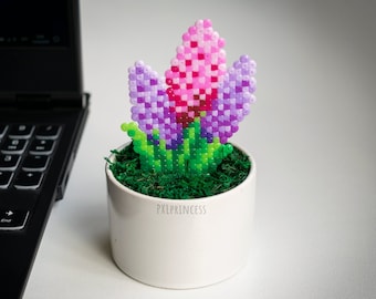 Może przedstawiać: Mały biały ceramiczny doniczek z zieloną podstawą z mchu i fioletowym i różowym kwiatem wykonanym z koralików perler. Doniczek ma napis "PXLPRINCESS" z przodu.