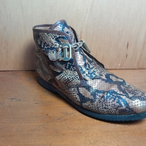 May include: Brown ankle boots with a snakeskin pattern and gold buckles. The boots have a lace-up closure and a rounded toe.