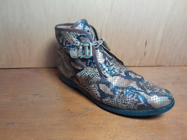 May include: Brown ankle boots with a snakeskin pattern and gold buckles. The boots have a lace-up closure and a rounded toe.