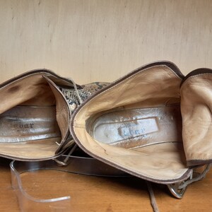 May include: A pair of brown leather ankle boots with a snakeskin pattern. The boots are unlaced and the inside of the boots is visible. The brand name "G.Bar" is visible on the inside of the left boot.