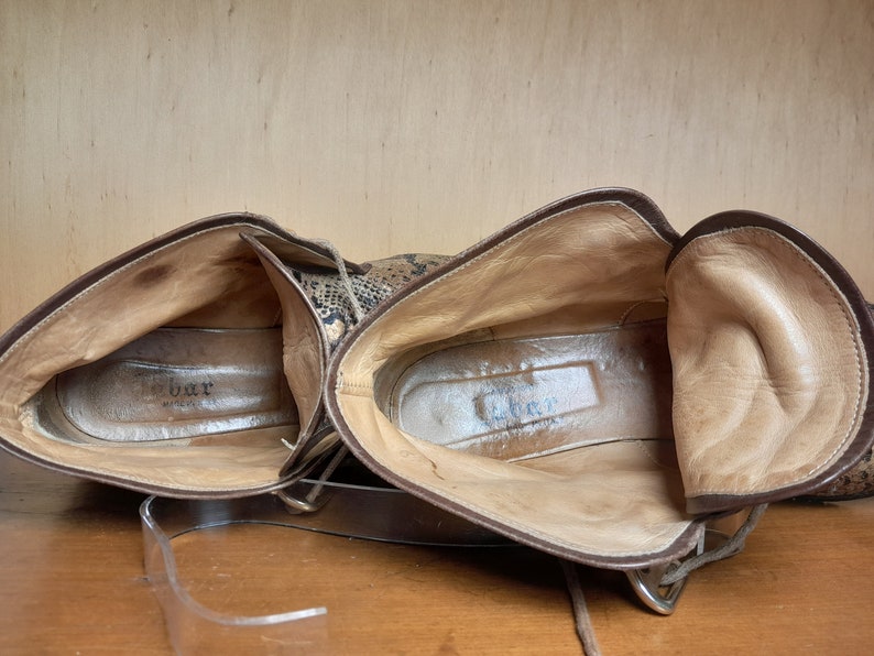 May include: A pair of brown leather ankle boots with a snakeskin pattern. The boots are unlaced and the inside of the boots is visible. The brand name "G.Bar" is visible on the inside of the left boot.