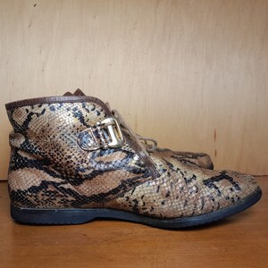 May include: Brown and gold snakeskin ankle boots with a buckle detail. The boots have a lace-up closure and a rounded toe.