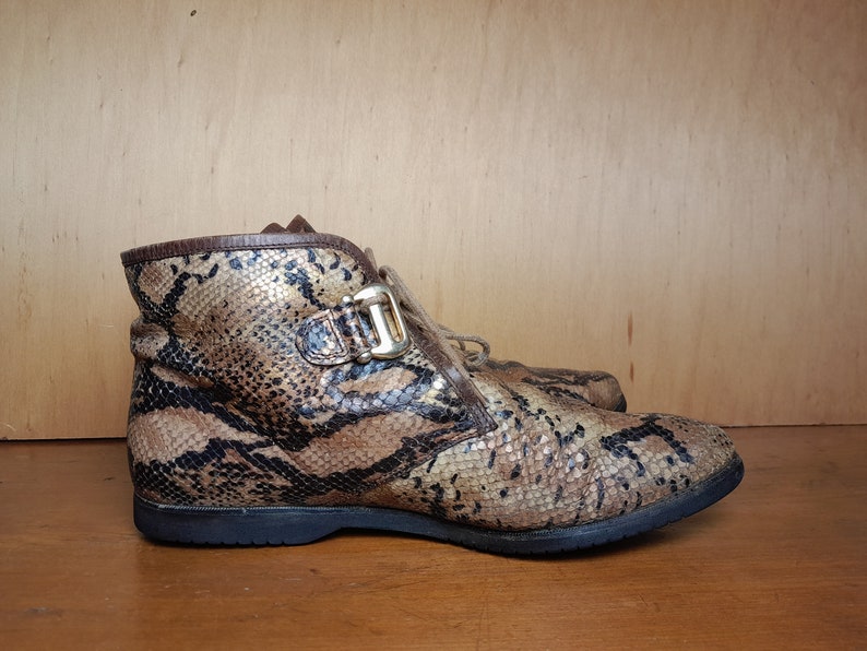 May include: Brown and gold snakeskin ankle boots with a buckle detail. The boots have a lace-up closure and a rounded toe.