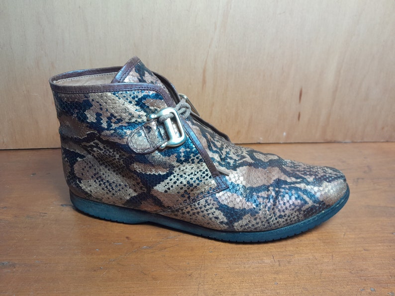 May include: Brown ankle boots with a snakeskin pattern and a gold buckle. The boots have a lace-up closure and a rounded toe.