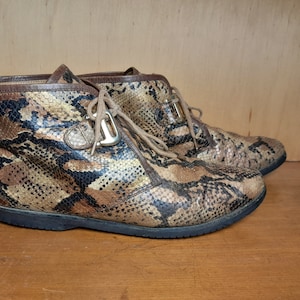 Pode incluir: Um par de botas de tornozelo de couro de cobra marrom e preto com cadarços e uma fivela dourada.