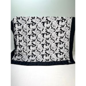May include: A black and white paisley patterned blanket. The blanket features a repeating design of white paisley shapes on a black background. The edges are trimmed with a solid black border. The blanket is folded and displayed on a white surface.