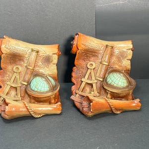 Könnte beinhalten: Zwei dekorative Resin-Tafeln mit einem Schriftrollen-Design, das eine Karte, ein Teleskop, einen Kompass und einen Globus zeigt. Die Tafeln sind in Braun-, Orange- und Türkistönen gehalten, mit dem Wort "Hispaniola".