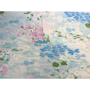 Puede incluir: Tela floral con fondo azul claro y blanco. La tela presenta racimos de margaritas rosas y blancas y flores azules. También se ven hojas y tallos verdes, creando un suave efecto acuarela.