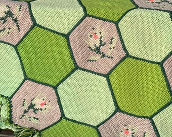 Könnte beinhalten: Eine handgefertigte Häkeldecke mit einem sechseckigen Muster in Grün- und Mauve-Tönen. Jedes Sechseck ist in einem dunkleren Grün umrandet, mit floralen Stickereien in der Mitte einiger. Die Decke hat einen gewellten Rand mit grünen Quasten.
