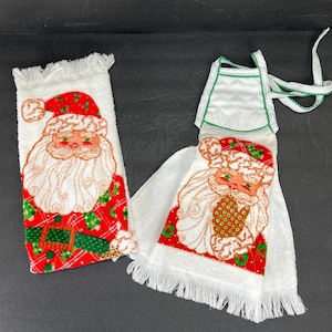 May include: Two Christmas-themed kitchen towels. One is a rectangular hand towel with a Santa Claus design, featuring a red hat and suit with green accents. The other is a white towel with a Santa Claus design and a green-trimmed apron top.