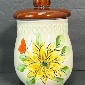 Op de afbeelding: Een witte keramische pot met een bruin deksel en een geschilderd bloemenontwerp. De pot heeft een gele bloem met groene bladeren en een rode vlinder.