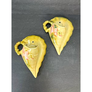 May include: Two yellow ceramic wall planters shaped like seashells with pink floral designs. The planters have a small hole at the top for drainage.