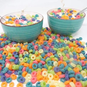 May include: Two teal bowls filled with colorful cereal rings and milk. The bowls are surrounded by a scattering of the cereal rings.