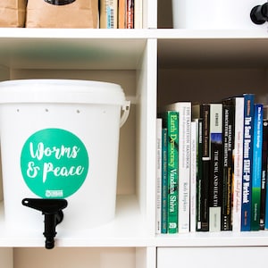 May include: A white composting bin with a black tap and the words "Worms & Peace" in teal. The bin is on a shelf with books about agriculture and sustainability.