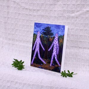 May include: A greeting card featuring an illustration of two purple figures with dotted patterns walking in a forest. The background shows trees and a blue sky. The card is on a white surface with green leaves.