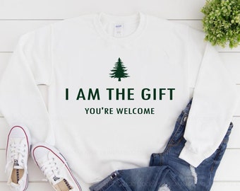 Op de afbeelding: Witte trui met een groene dennenboom afbeelding en de tekst "I AM THE GIFT YOU'RE WELCOME".