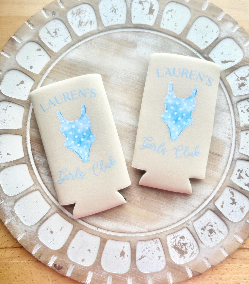 May include: Two beige can coolers with a blue and white polka dot swimsuit design. The text "Lauren's Girls Club" is printed on each cooler.