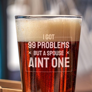 May include: A pint glass with a brown beverage and a white inscription that reads "I got 99 problems but a spouse ain't one". The glass is sitting on a wooden surface next to a brown glass bottle.