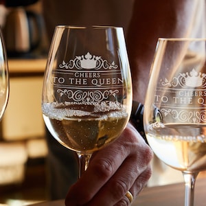 May include: Two clear wine glasses with the text "Cheers to the Queen" etched on the side. Each glass is filled with white wine.