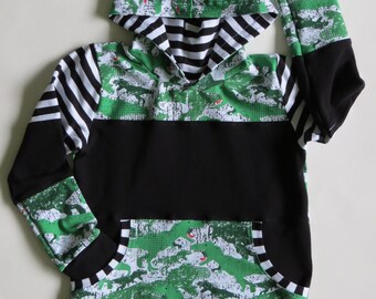 May include: Black and green hooded sweatshirt with a camouflage print and a black and white striped inner lining. The sweatshirt has a large pocket on the front and a small label that says "The Handmade Market".