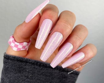 May include: Close-up of a hand with long, light pink glitter nails. The nails are long and almond shaped. The hand is wearing a pink and white checkered ring.