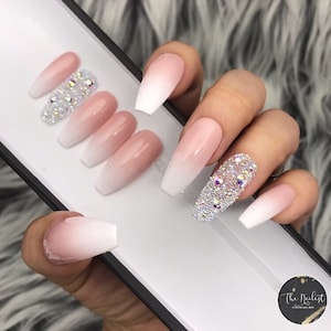 May include: Set of long, coffin-shaped, ombre press-on nails with a pink to white gradient. One nail features a large cluster of clear rhinestones.