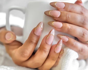 May include: Close-up of hands with almond-shaped nails painted a light pink with a gold accent design. The nails are long and pointed.