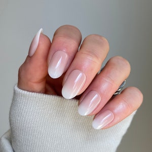 May include: Close-up of a hand with five oval-shaped nails painted with a white to light pink ombre nail polish.
