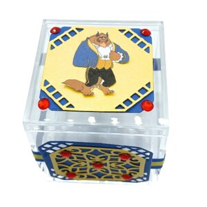 May include: A clear acrylic box with a yellow top featuring a cartoon image of the Beast from Beauty and the Beast. The box has blue and gold decorative accents and red embellishments. The box is square and has a hinged lid.