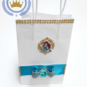 Może przedstawiać: Biała torba na prezent ze złotym obszyciem z dżet&oacute;w i turkusową kokardą. Zawiera złote zdjęcie Arielki z Małej Syrenki. Torba ma białe uchwyty ze sznurka i tekst "Royal Baby Baby Us" w okrągłym logo.