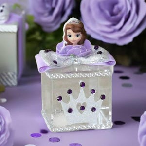 May include: A clear, square box with a princess figurine on top, decorated with a silver bow and a white crown with purple gems. The box is surrounded by purple roses and confetti on a purple surface.