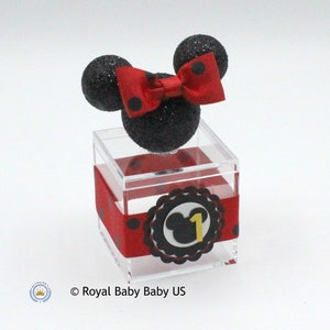 K&ouml;nnte beinhalten: Eine transparente Plastikbox in W&uuml;rfelform mit einem roten und schwarzen gepunkteten Band darum. Die Box hat einen schwarzen und roten Mickey-Maus-Kopf mit einer roten Schleife oben drauf. Die Box hat einen schwarzen und wei&szlig;en Kreis mit einem Mickey-Maus-Kopf und der Zahl 1 in Gelb darauf.