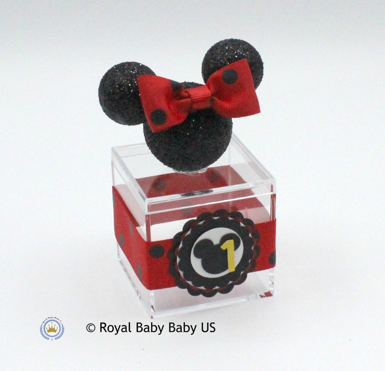 K&ouml;nnte beinhalten: Eine transparente Plastikbox in W&uuml;rfelform mit einem roten und schwarzen gepunkteten Band darum. Die Box hat einen schwarzen und roten Mickey-Maus-Kopf mit einer roten Schleife oben drauf. Die Box hat einen schwarzen und wei&szlig;en Kreis mit einem Mickey-Maus-Kopf und der Zahl 1 in Gelb darauf.
