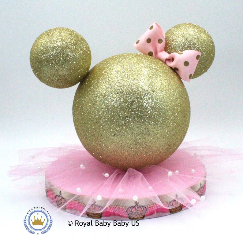 May include: Gold glitter Mickey Mouse ears centerpiece with a pink polka dot bow and a pink tulle skirt with pearl accents. The base is decorated with a pink and gold crown ribbon.