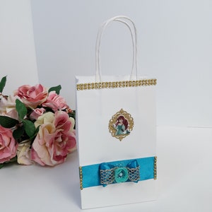 Może przedstawiać: Biała torba na prezent ze złotym wykończeniem i turkusową wstążką. Torba posiada oprawiony obrazek postaci z kresk&oacute;wki. Do wstążki przymocowana jest turkusowa kokarda z perłowym środkiem.