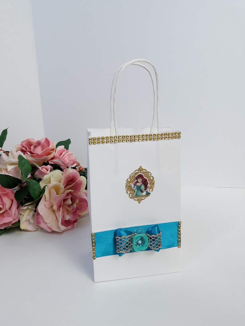 Może przedstawiać: Biała torba na prezent ze złotym wykończeniem i turkusową wstążką. Torba posiada oprawiony obrazek postaci z kresk&oacute;wki. Do wstążki przymocowana jest turkusowa kokarda z perłowym środkiem.