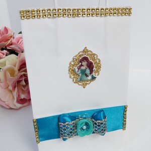 Może przedstawiać: Biała torba na prezent ze złotym obszyciem z dżet&oacute;w i turkusową kokardą. Zawiera złotą, oprawioną grafikę postaci z kresk&oacute;wki z rudymi włosami i turkusową sukienką. Torba ma białe papierowe uchwyty.