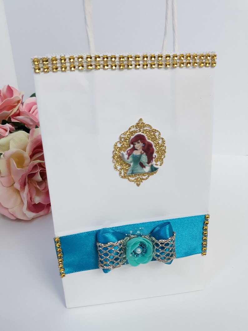 Może przedstawiać: Biała torba na prezent ze złotym obszyciem z dżet&oacute;w i turkusową kokardą. Zawiera złotą, oprawioną grafikę postaci z kresk&oacute;wki z rudymi włosami i turkusową sukienką. Torba ma białe papierowe uchwyty.